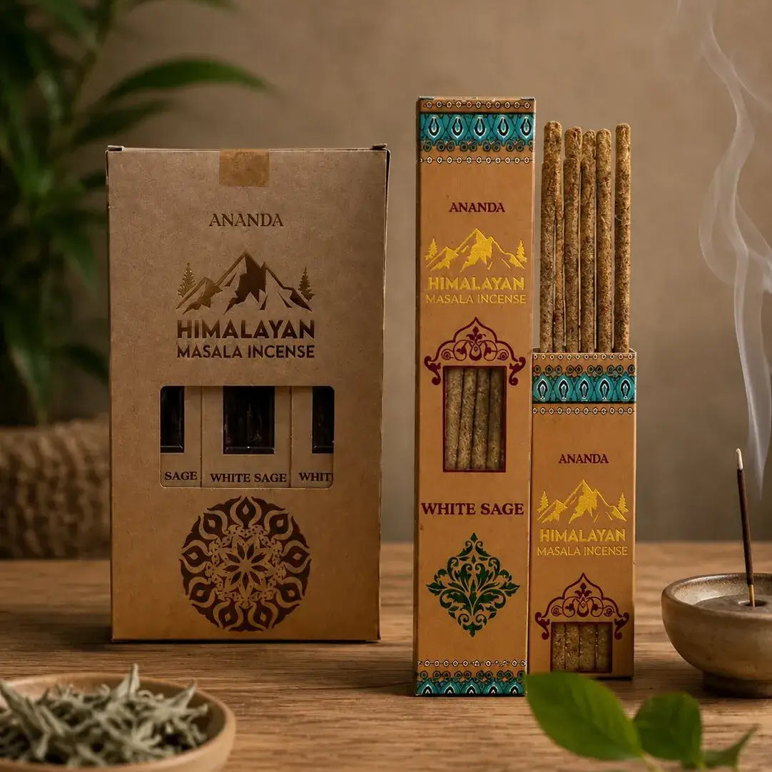 Three boxes of Ananda Himalayan Masala Incense on a wooden table, with one open box showing incense sticks, a lit incense stick smoking, a small bowl of dried herbs, and green leaves in a styled setting.